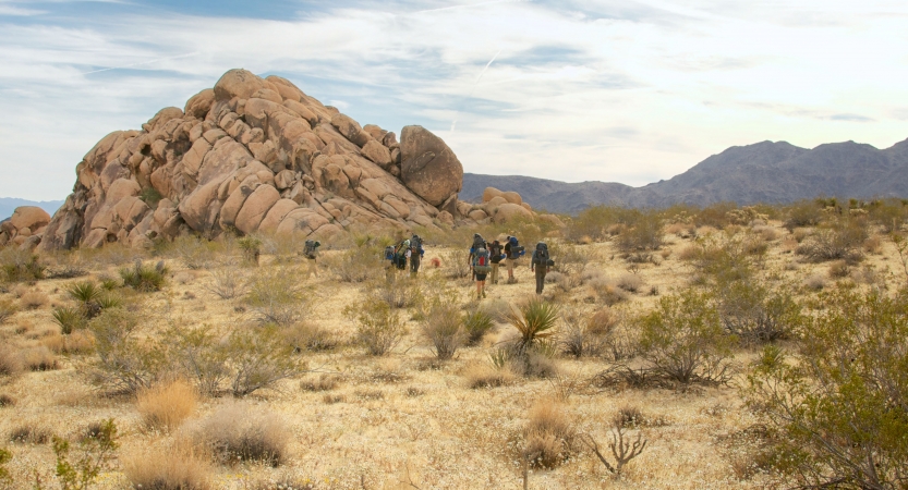 From a distance, a group of backpackers make their way through a desert landscape toward a large rock formation. 